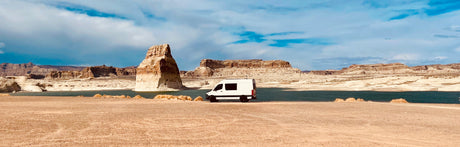 RV rental van on a desert lake shore, with a towering rock formation and distant mesas under a blue sky.