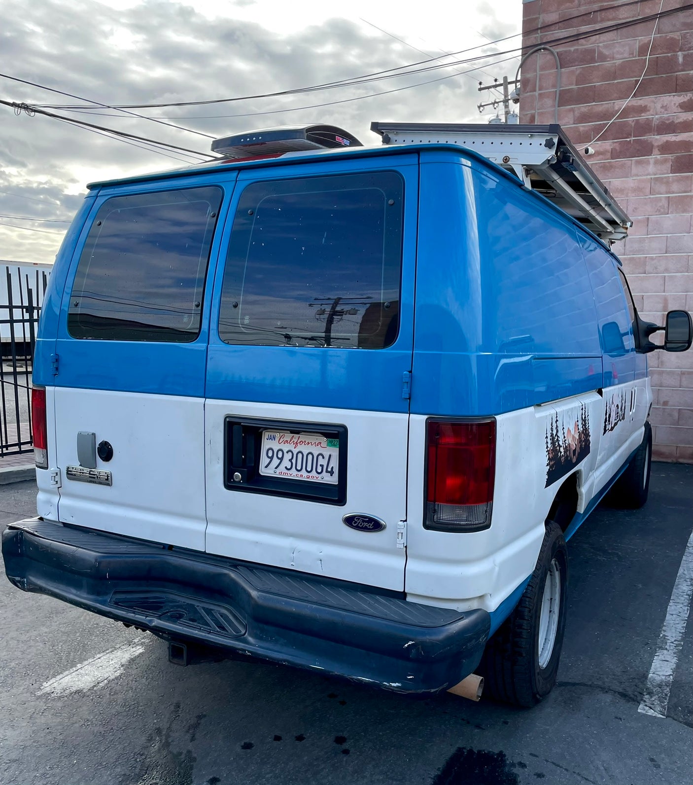 RV rental: Rear view of a custom blue and white Ford Econoline camper van with a roof rack, solar panels, and nature decals.