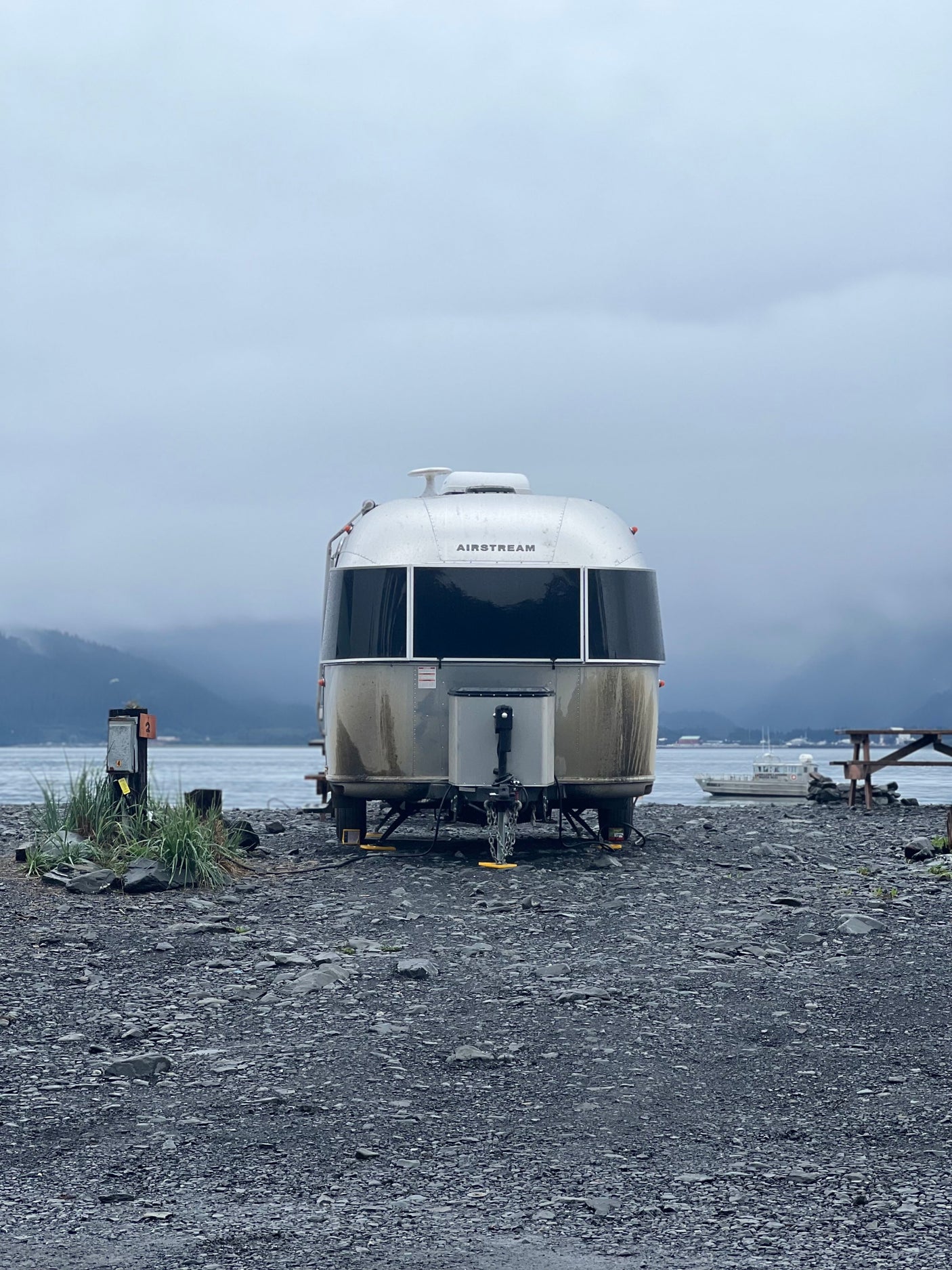 A large Airstream RV rental is parked on a grey rocky shore, facing a misty body of water and mountains. Overcast skies.
