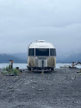 A large Airstream RV rental is parked on a grey rocky shore, facing a misty body of water and mountains. Overcast skies.