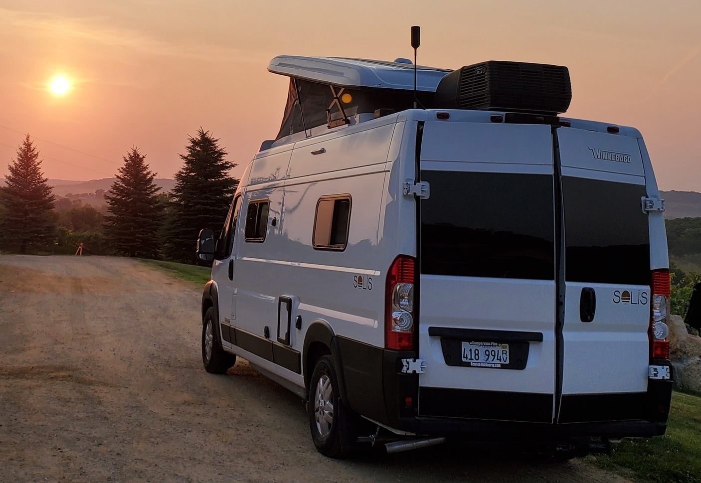 A white Winnebago Solis rv rental at sunset, pop-top open, on a scenic dirt road with distant hills.