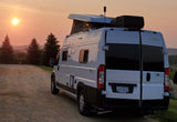 A white Winnebago Solis rv rental at sunset, pop-top open, on a scenic dirt road with distant hills.