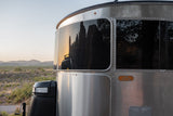 Close-up of a silver Airstream rv rental, its front windows reflecting jagged desert peaks at sunset.