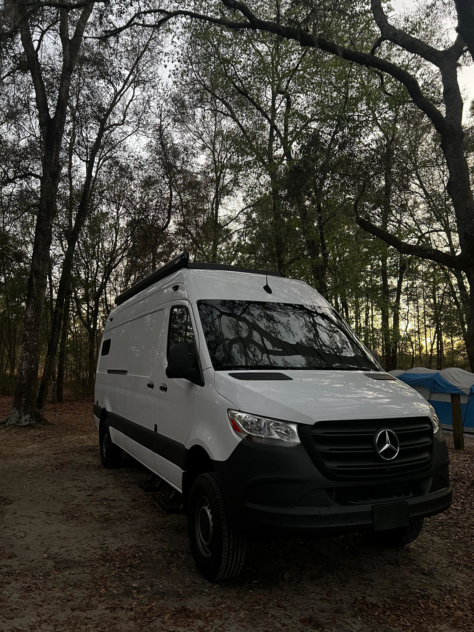 White Mercedes Sprinter rv rental camper van at a wooded campsite with a tent, ready for your outdoor adventure.