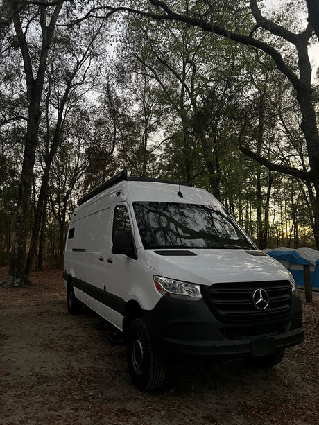 White Mercedes Sprinter rv rental camper van at a wooded campsite with a tent, ready for your outdoor adventure.