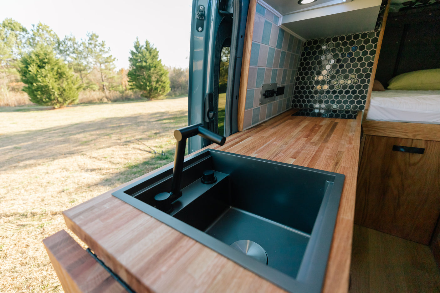 Inside a custom RV rental: modern kitchen with black sink, wood counter, stylish tile backsplash, and bed visible.