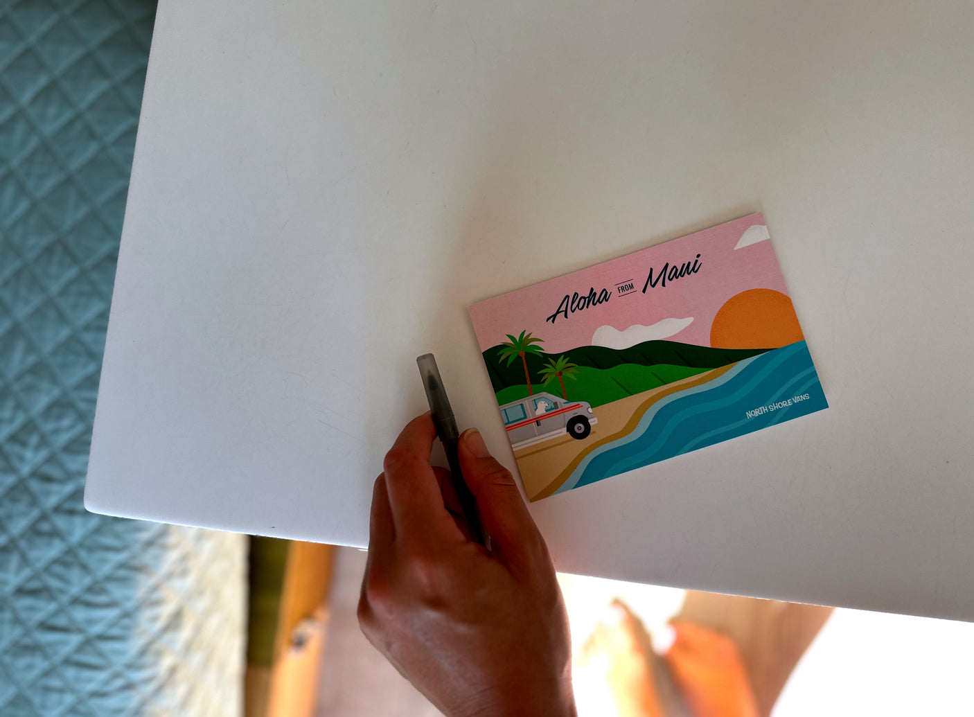 A hand holds a pen on a white surface next to an "Aloha From Maui" postcard, promoting North Shore Vans RV rental.