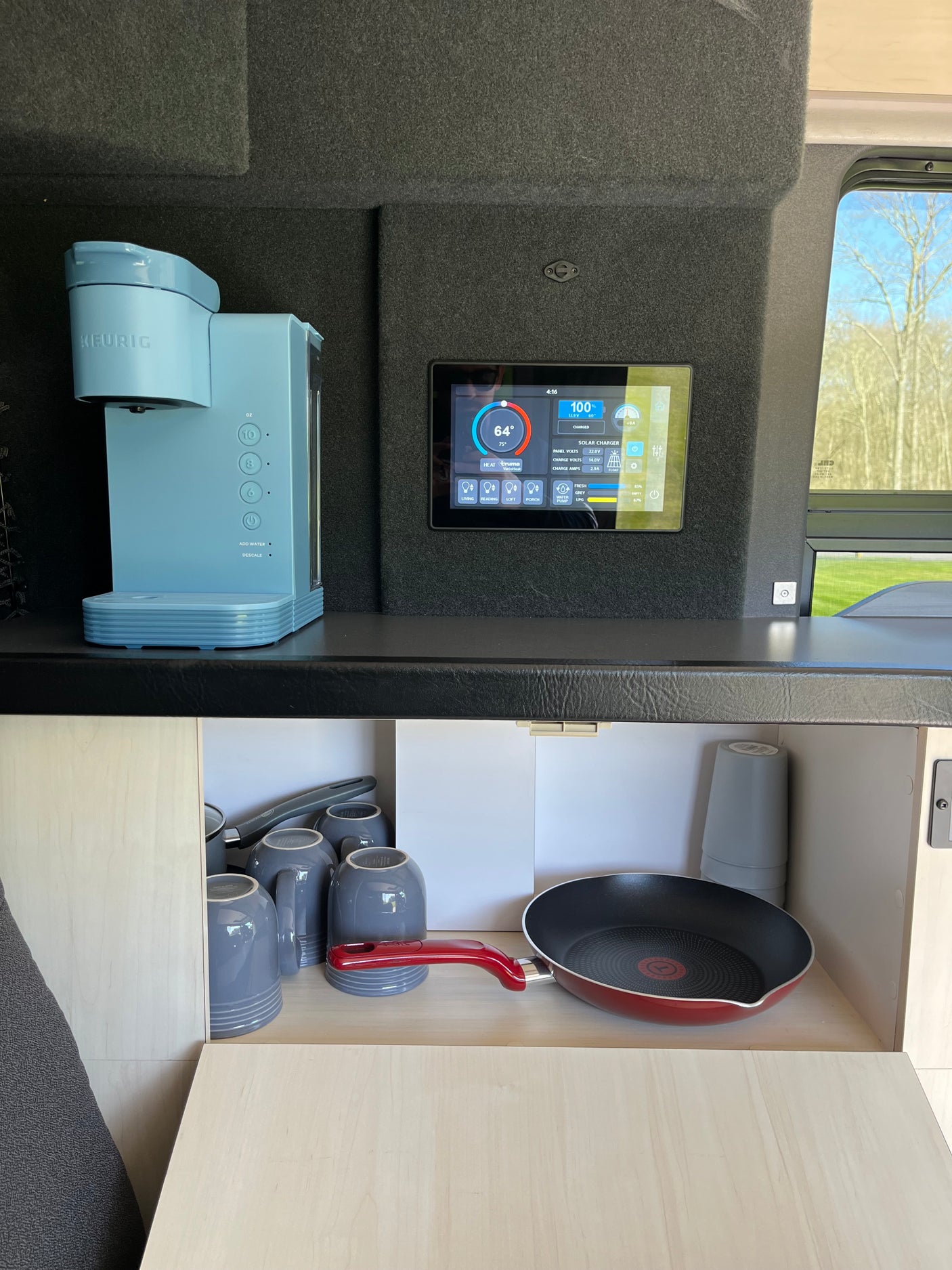 Inside an RV rental: a light blue Keurig coffee maker, a smart control panel for power and water, plus kitchenware like mugs and a frying pan.