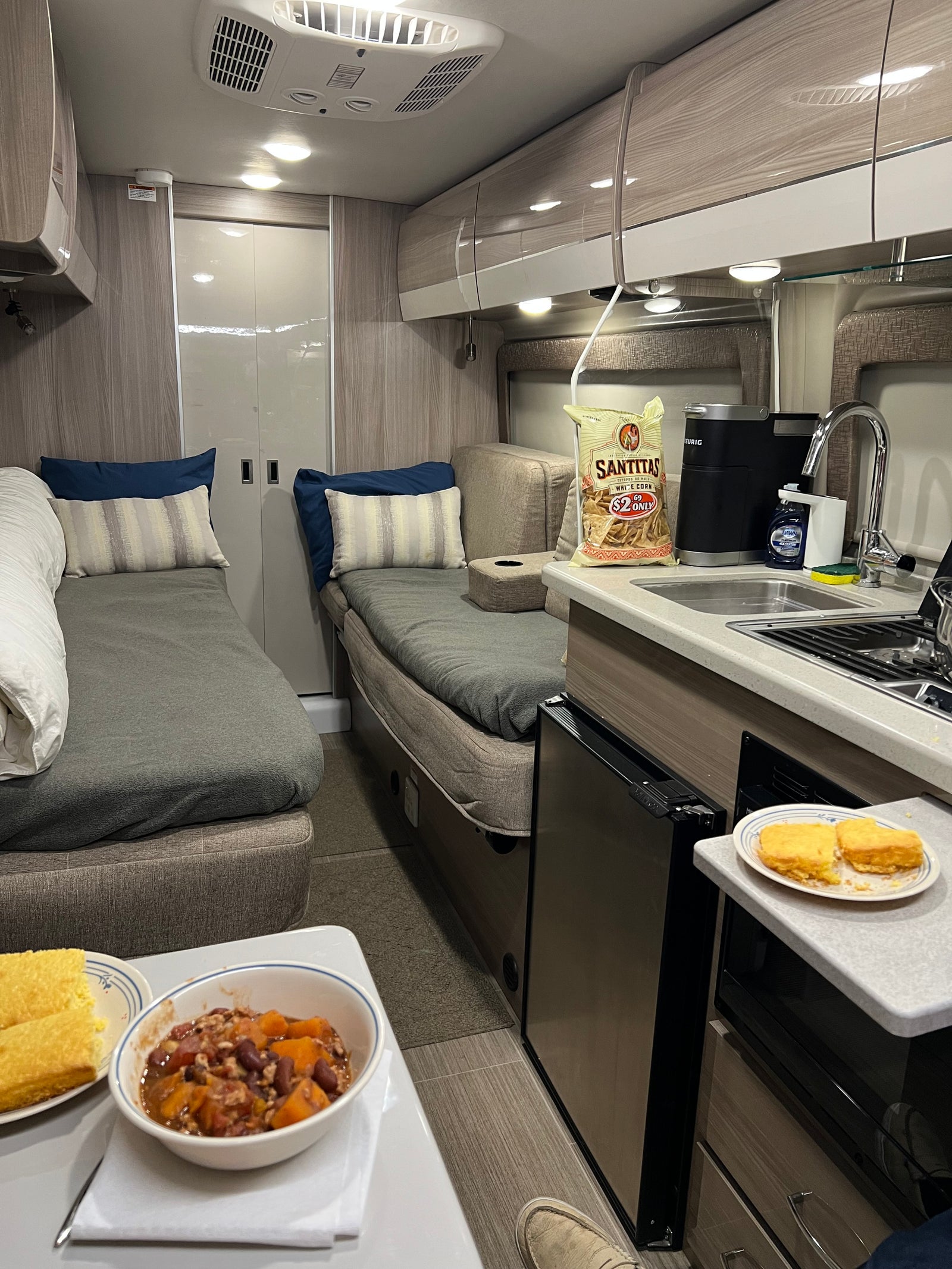 Inside a cozy RV rental: two sleeping/lounge areas, a kitchenette with coffee and snacks, plus a warm meal ready.