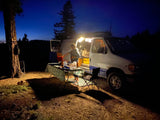 Nighttime outdoor cooking with an RV rental setup. A person prepares food by a lighted camper van and table under a starry sky.