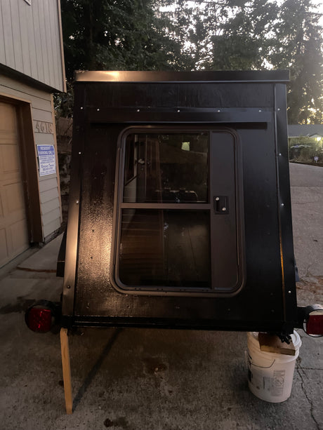 Rear view of a compact black custom-built camper with a windowed door, propped on blocks, ready for **rv rental**.