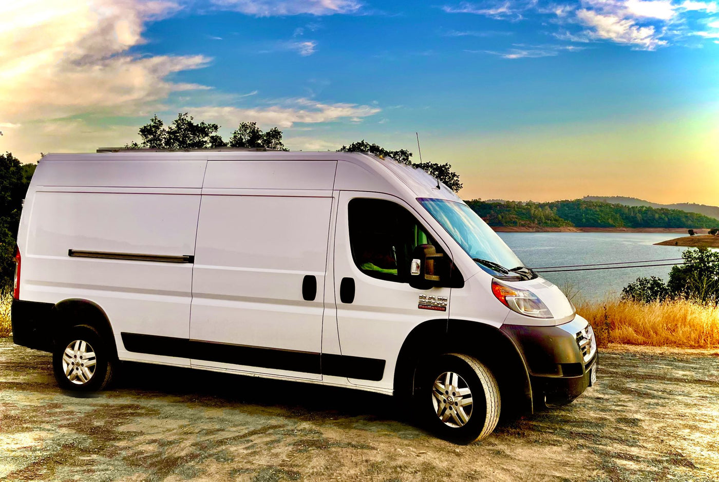  kitchen and nature views visible.|RV rental: a white Ram ProMaster camper van parked by a scenic lake under a colorful sky.|This white Ram ProMaster camper van — photo 1