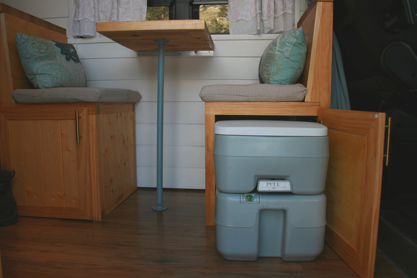 Compact RV rental interior: wooden seating, cushions, a table, and a grey portable toilet accessible via an open cabinet.