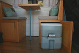 Compact RV rental interior: wooden seating, cushions, a table, and a grey portable toilet accessible via an open cabinet.