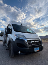 A white Ram RV rental van, parked on a gravel lot under a dramatic cloudy sky, with mountains in the background.
