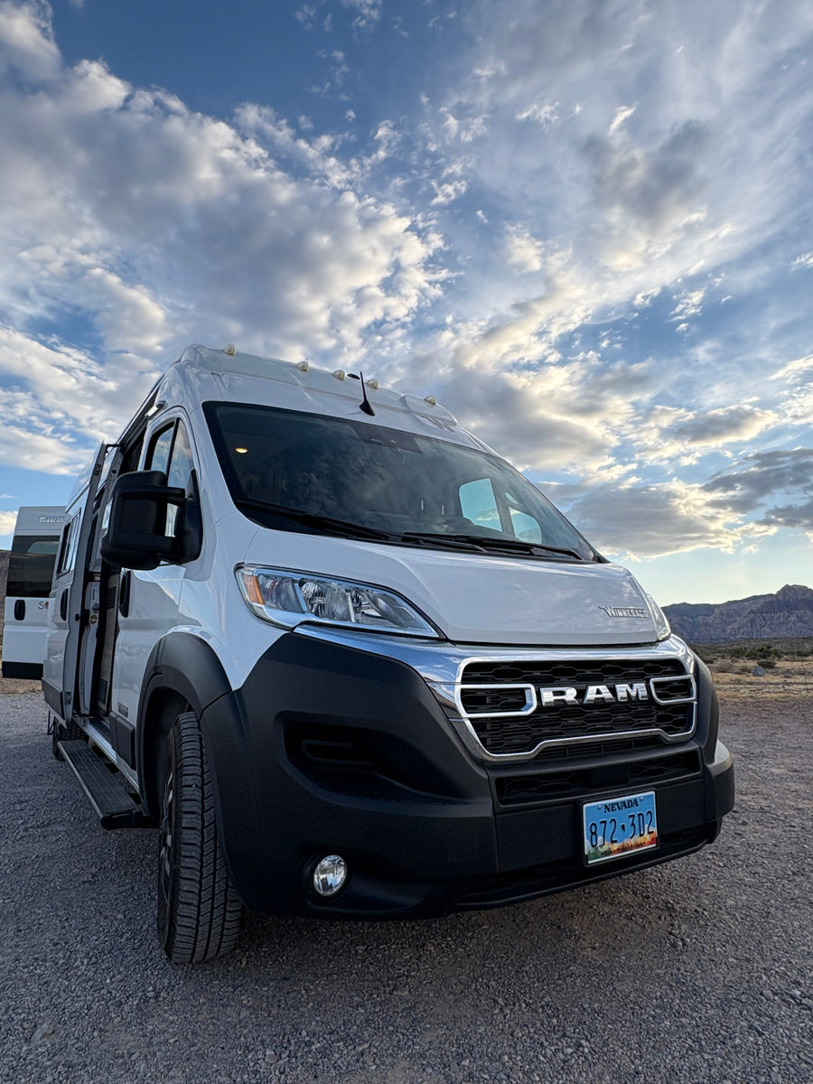 A white Ram RV rental van, parked on a gravel lot under a dramatic cloudy sky, with mountains in the background.