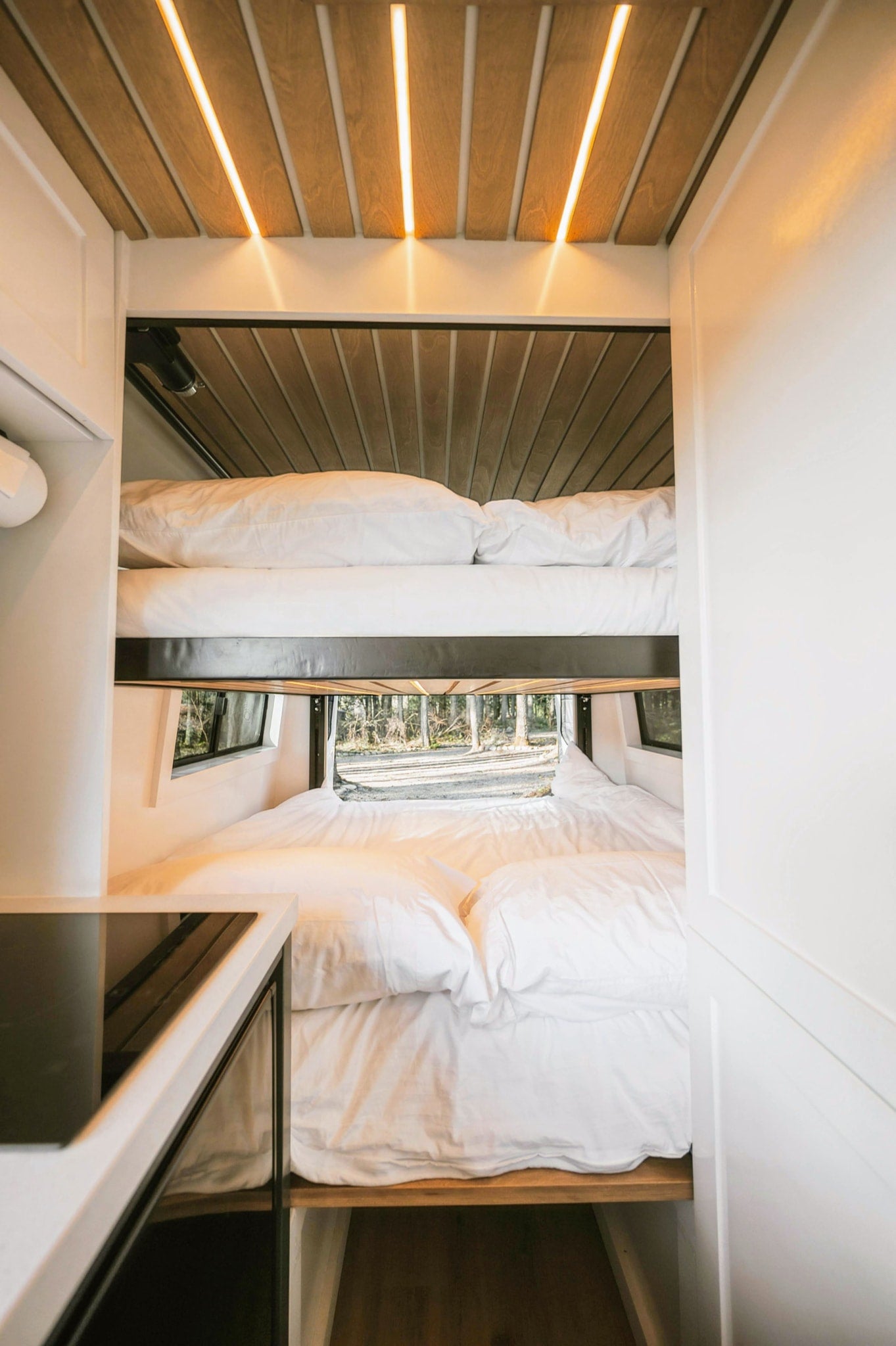 Inside a modern RV rental: stacked bunk beds with white linens, ambient wood ceiling lighting, and a window view of a serene forest. — photo 4