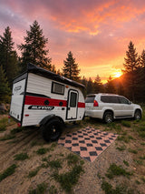 RV rental: A Sunray Sport teardrop camper & Lexus GX SUV parked amidst pines at a stunning sunset. Checkerboard mat set up for camping.