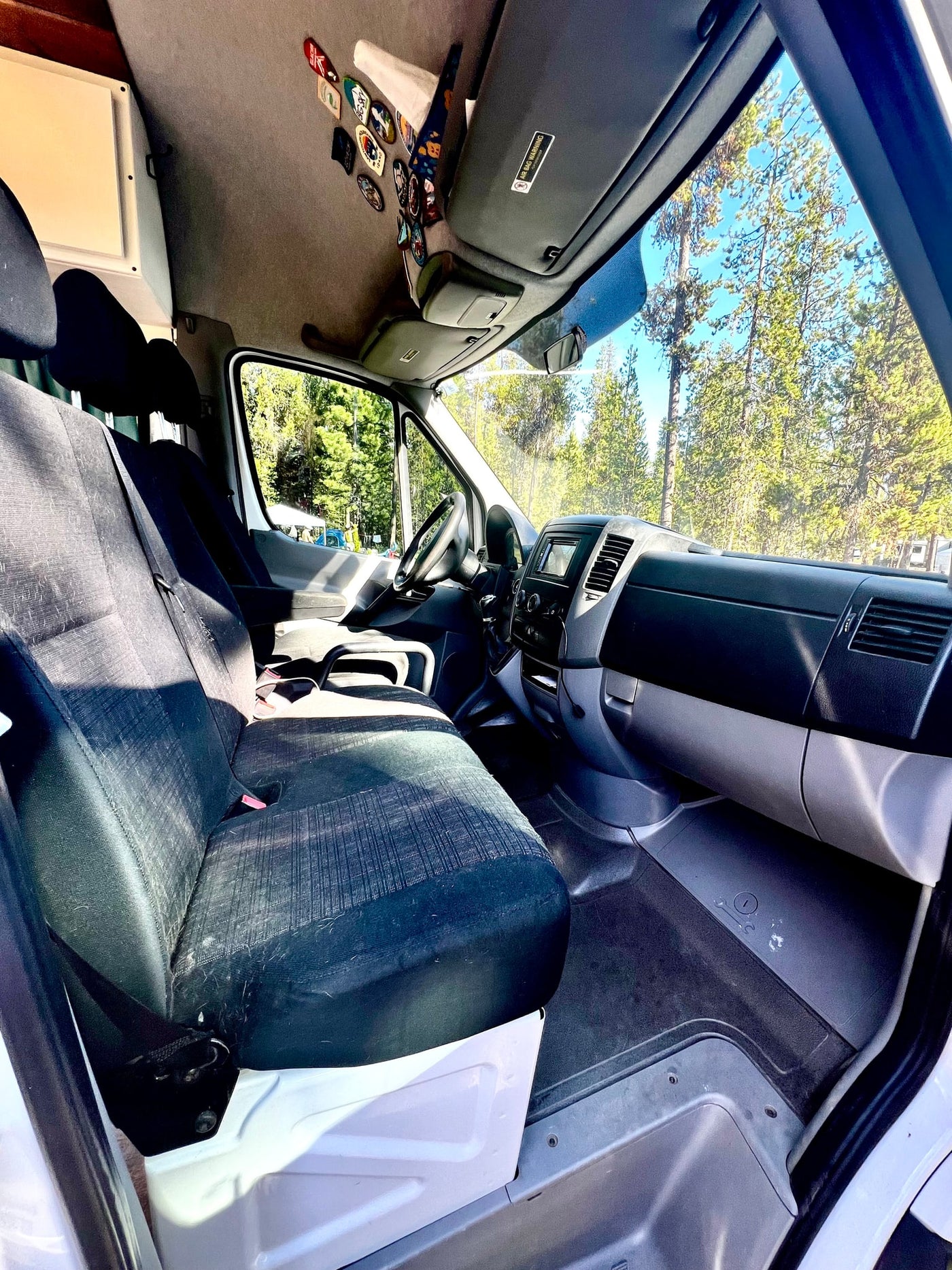 An RV rental's front cabin: dark fabric seats, dash, and windshield view of sunny forest. — photo 11