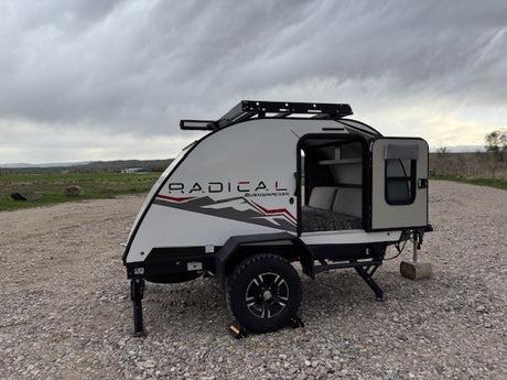 Explore the outdoors with this Radical Bushwhacker teardrop RV rental. Parked on gravel, its open door shows a cozy interior.