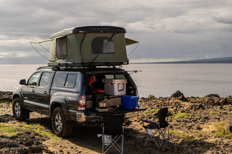  parked by the ocean at sunset. A unique RV rental option for adventure travel.|Overland RV rental: A dark pickup truck with a rooftop tent and tailgate camp kitchen by a vast body of water.|Experience RV rental adventure: A Tacoma truck with rooftop tent ready for beachside camping in Hawaii at sunset.|Rugged Toyota Tacoma truck with a rooftop tent tackles a rocky Hawaiian trail. An ideal adventure vehicle — photo 2