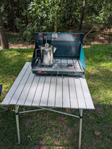 Coleman stove with coffee pot on a camping table at an RV rental campsite, ready for adventure.