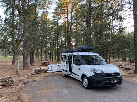 White Ram ProMaster City RV rental parked at a forest campsite with an open door, roof box, and rear cooler.