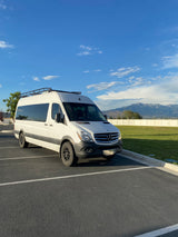 A white Mercedes Sprinter camper van with a roof rack, suitable for rv rental, parked in a lot with mountains and sky visible.