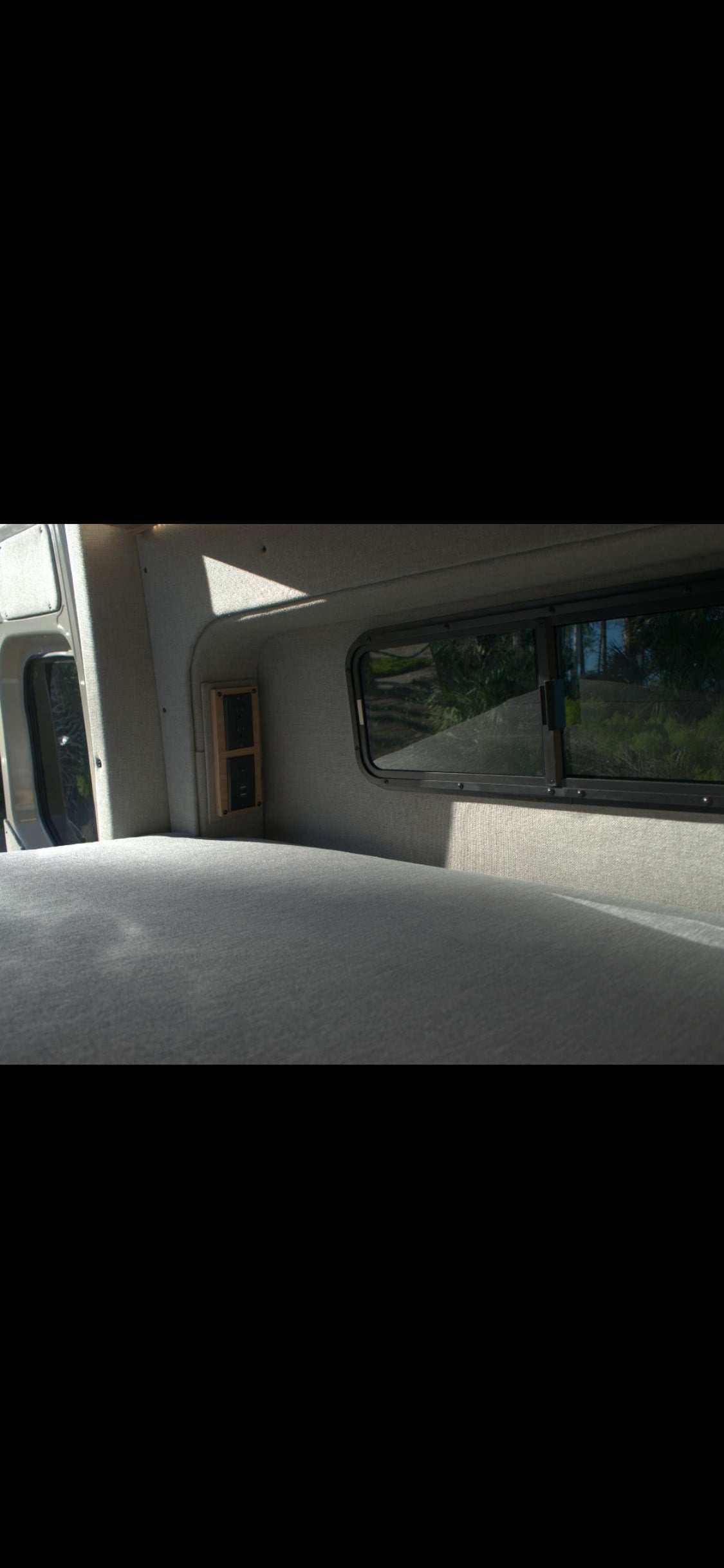 RV rental's cozy interior: grey bed, window with green outdoor view, and wall-mounted power outlets.