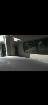 RV rental's cozy interior: grey bed, window with green outdoor view, and wall-mounted power outlets.