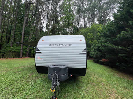 Gray Sun Lite Sport travel trailer RV rental parked in a lush, green campsite, surrounded by tall trees.