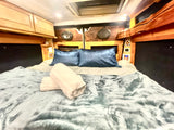Cozy RV rental bed with blue satin pillows, fluffy blanket, and rolled towels. Wooden paneling and cabinets line the walls.