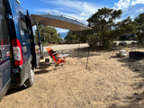 Relaxing at a dirt campsite with an RV rental. A blue camper van with an attached awning shades a person at a table.