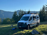 White Mercedes Sprinter RV rental under clear blue skies, parked in a scenic mountain meadow for off-grid adventure.