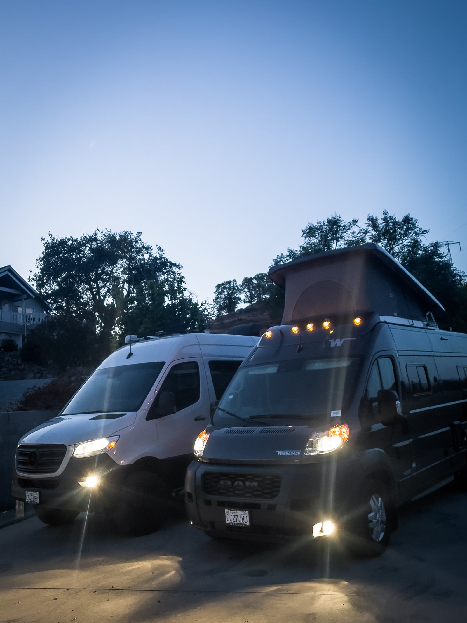 Two modern rv rental camper vans, a white Mercedes Sprinter and a dark Winnebago with a pop-top, are parked at dusk.
