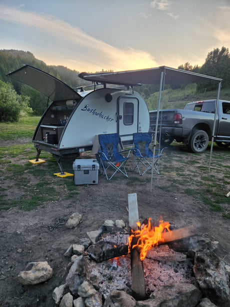Cozy rv rental setup: Bushwhacker trailer, chairs, and a blazing campfire at sunset. Perfect outdoor trip.