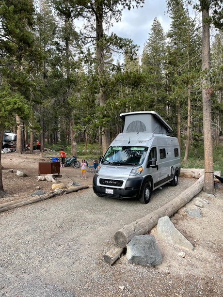 RV rental camping: Silver Winnebago camper van at a rustic site with people & bikes.