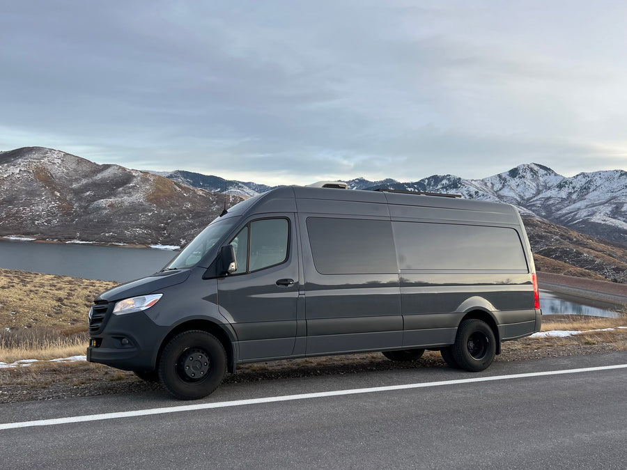 A dark grey Sprinter van, ideal for rv rental, parked by a roadside with a lake and snowy mountains in the background.