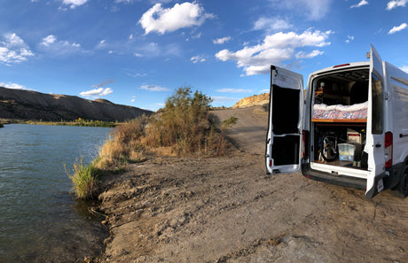 RV rental campervan at a scenic river spot, rear doors open revealing the cozy bed, bike & gear inside, ideal for camping.