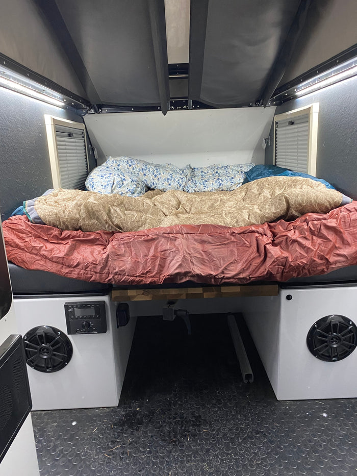  amidst pine trees at dusk.|Overland trailer (like an RV rental) & Lexus SUV with two happy dogs camping in pine trees. Adventure ready setup!|Silver SUV with rooftop tent towing an overland RV rental trailer at a peaceful forest campsite.|Cozy rv rental sleeping area features a bed with layered bedding