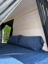 Bright RV rental interior: cozy bed with blue bedding, light wood walls, ambient LED, and lush trees outside.