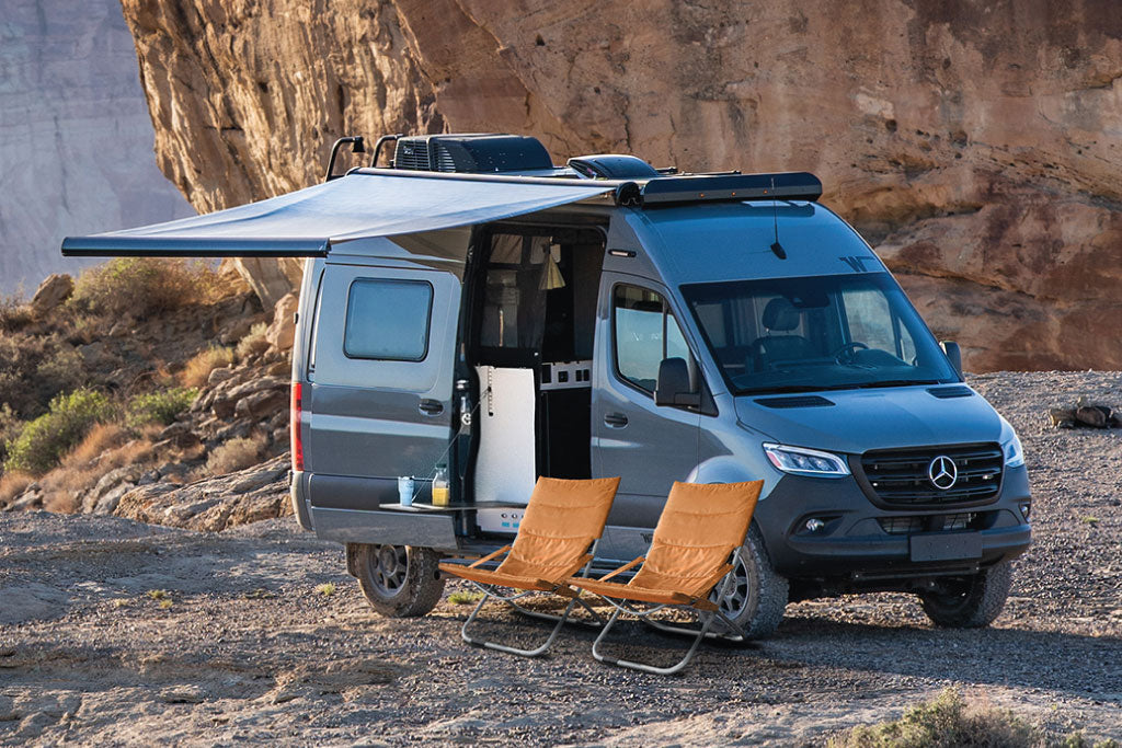 Grey Mercedes Sprinter camper van rv rental with awning & chairs set up for outdoor relaxation in a desert landscape.|A white custom camper van illuminates the night with bright LED and ground lights