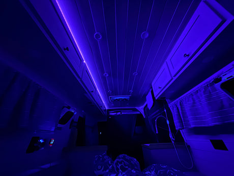 Dark blue-lit interior of an RV rental with overhead cabinets, slatted ceiling, and control panels for a modern campervan experience.