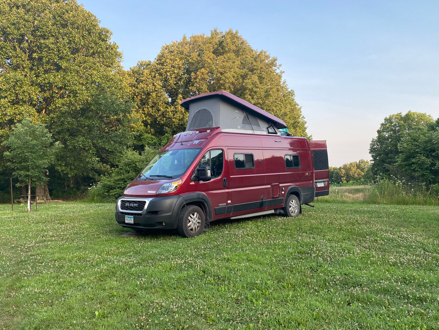 Red rv rental camper van with pop-top and open back doors, parked in a green field with trees. Ready for camping!