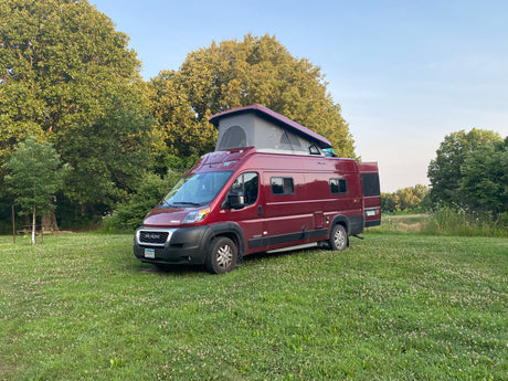 Red rv rental camper van with pop-top and open back doors, parked in a green field with trees. Ready for camping!