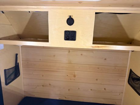 Inside an RV rental: light wood interior with overhead storage, mesh pockets, and multi-function power panel for charging.