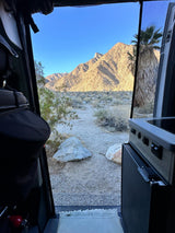 RV rental adventure: Desert mountains bathed in light, a dusty trail, and a palm are framed by the camper's open door.