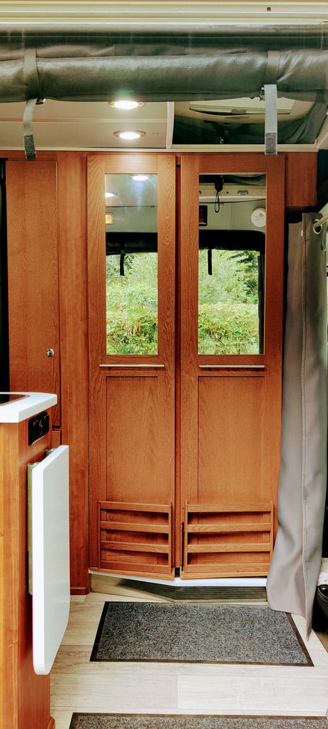 Inside an RV rental: rich wooden rear doors with windows framing lush nature. Practical interior features a kitchen area and lighting.