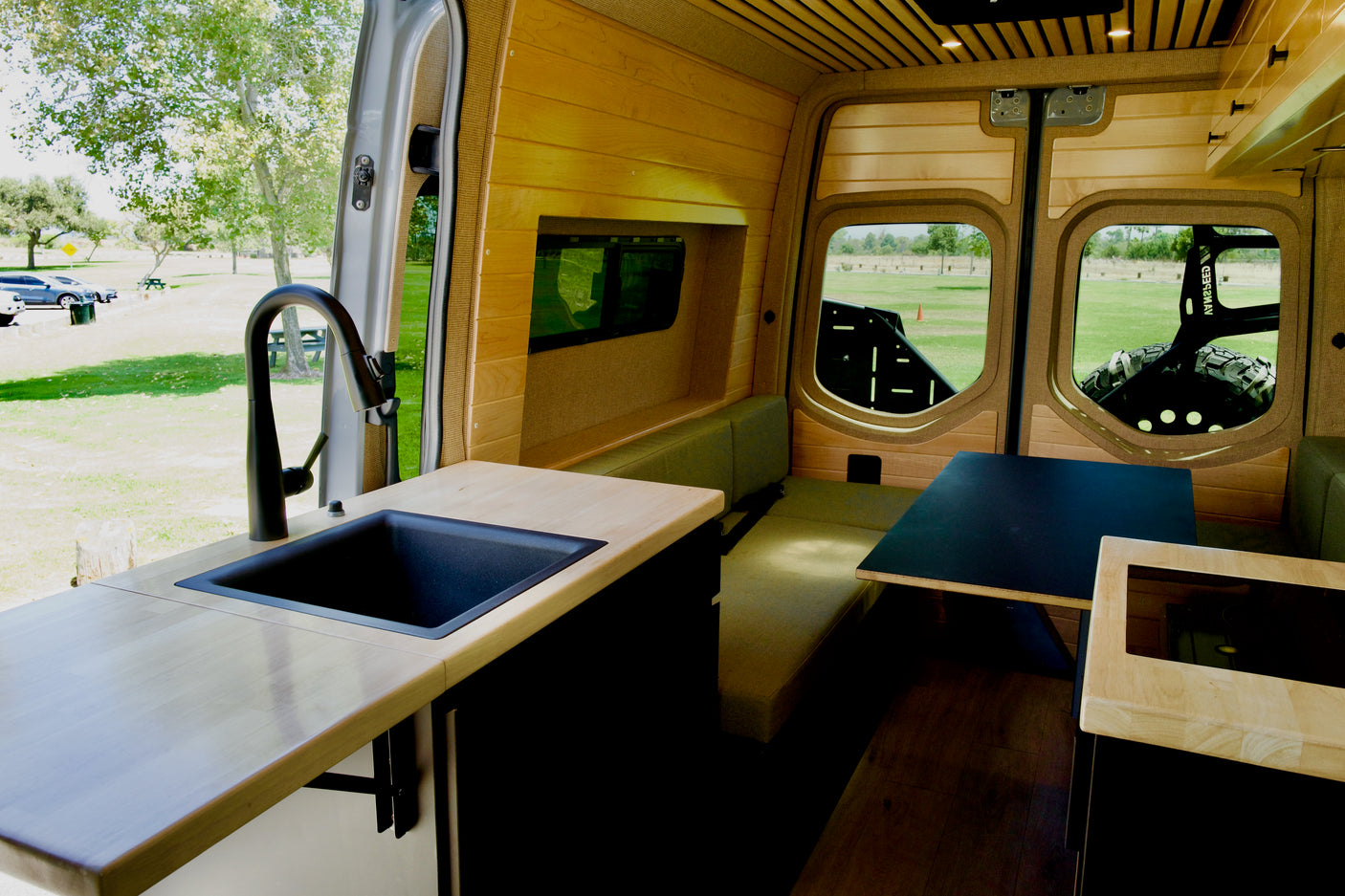 Inside a stylish rv rental camper van: light wood interior, kitchen with black sink/faucet, dining area, and scenic park view.