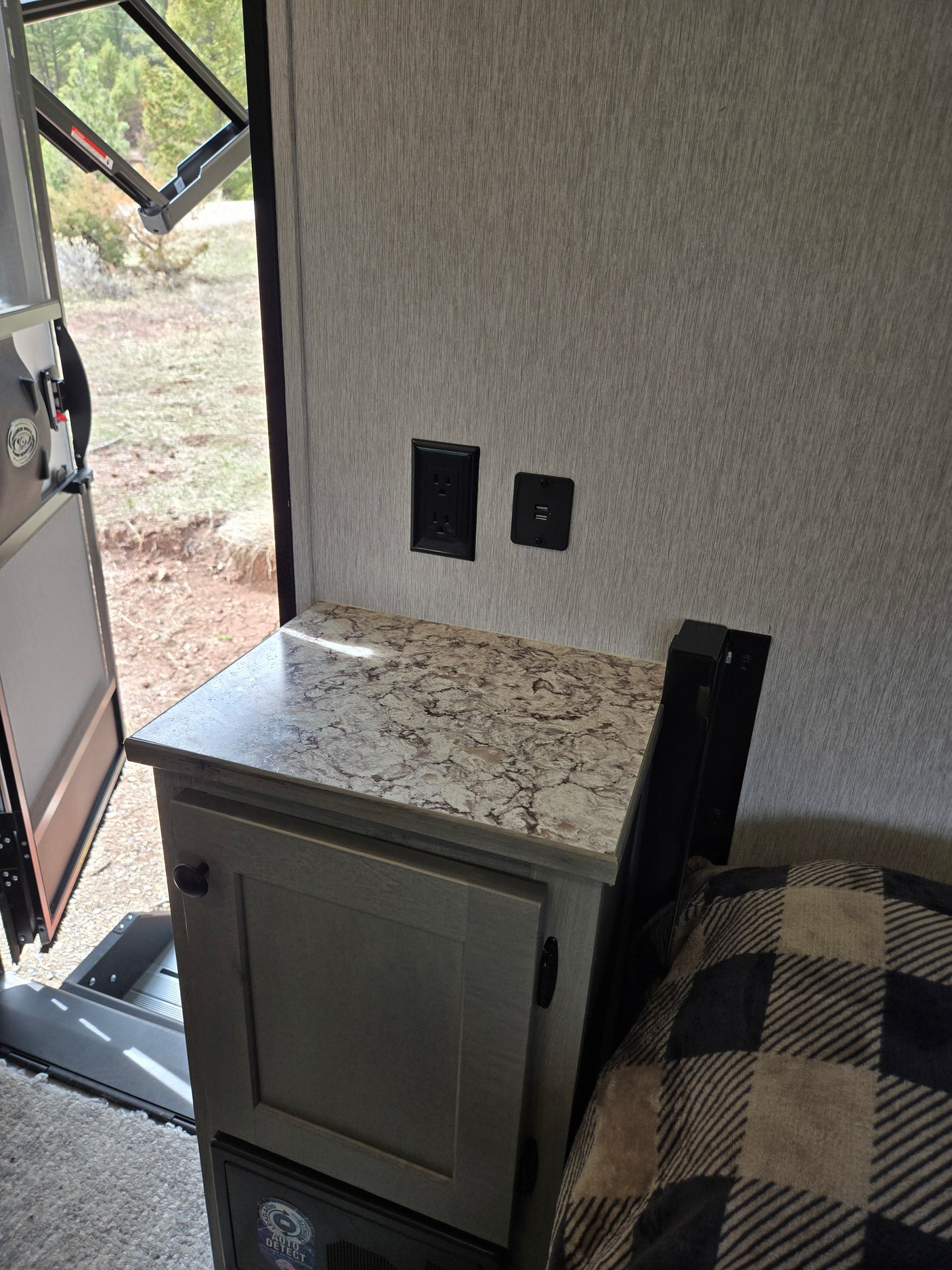 RV rental interior: nightstand with power outlets, open door to trees, and a cozy plaid blanket on the bed.